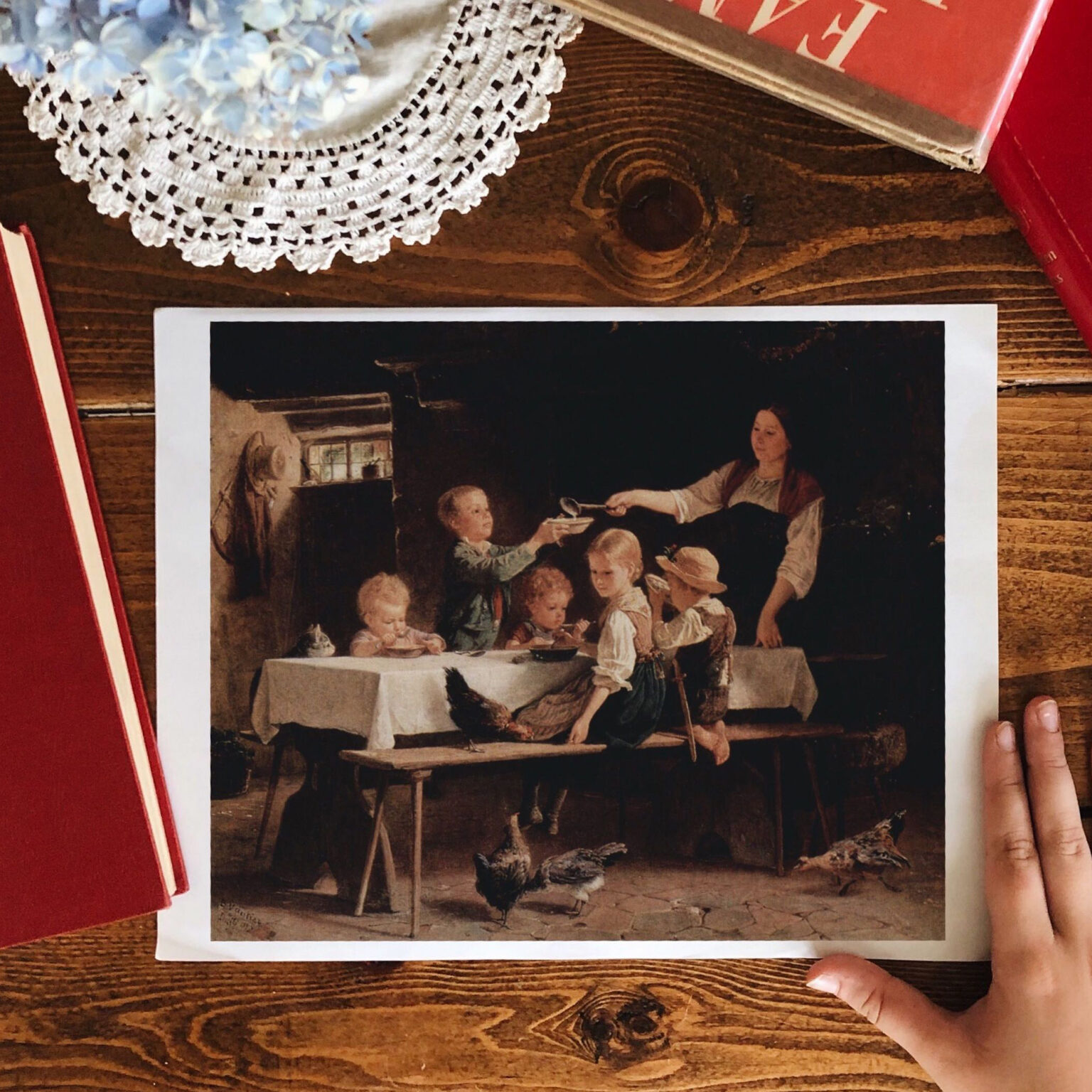 Printed reproduction of a classical family scene with children at a table, placed on a wooden table beside a crocheted doily and a red book on the edge of the frame.