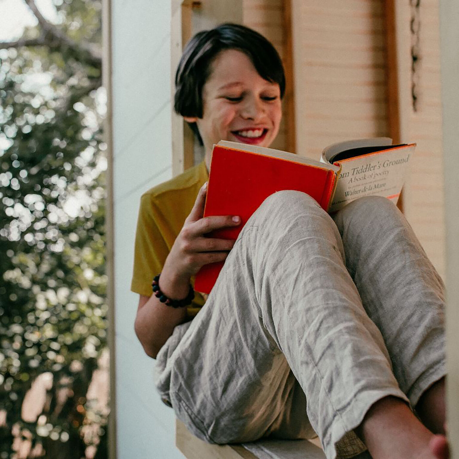 Person smiling while reading a bright red book outdoors, sitting on a railing or ledge with knees up, casual striped pants visible, blurred greenery in the background