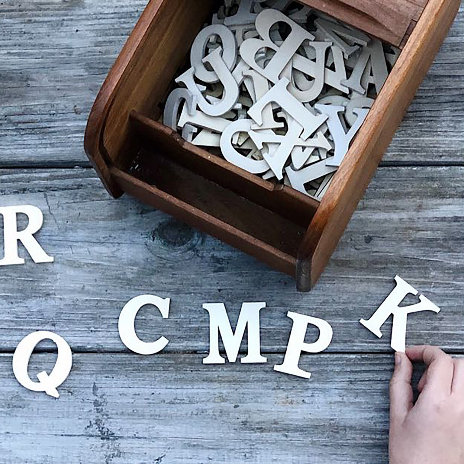 White uppercase letter cutouts scattered inside a wooden box on a weathered wooden table, with a hand reaching to pick one up.