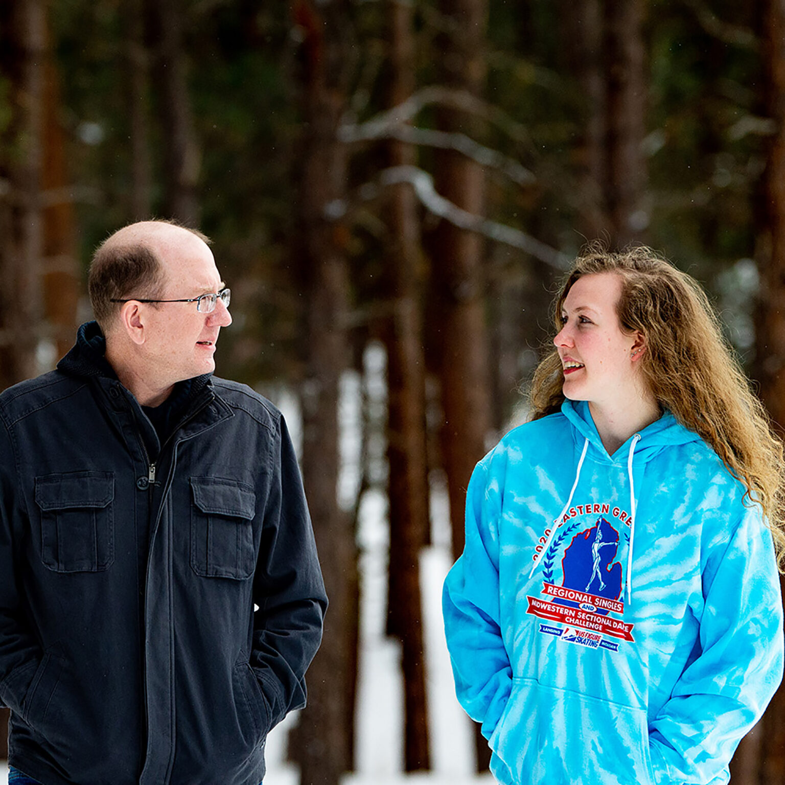 Man in a black jacket talks with a woman in a blue tie-dye hoodie outdoors in a snowy forest.