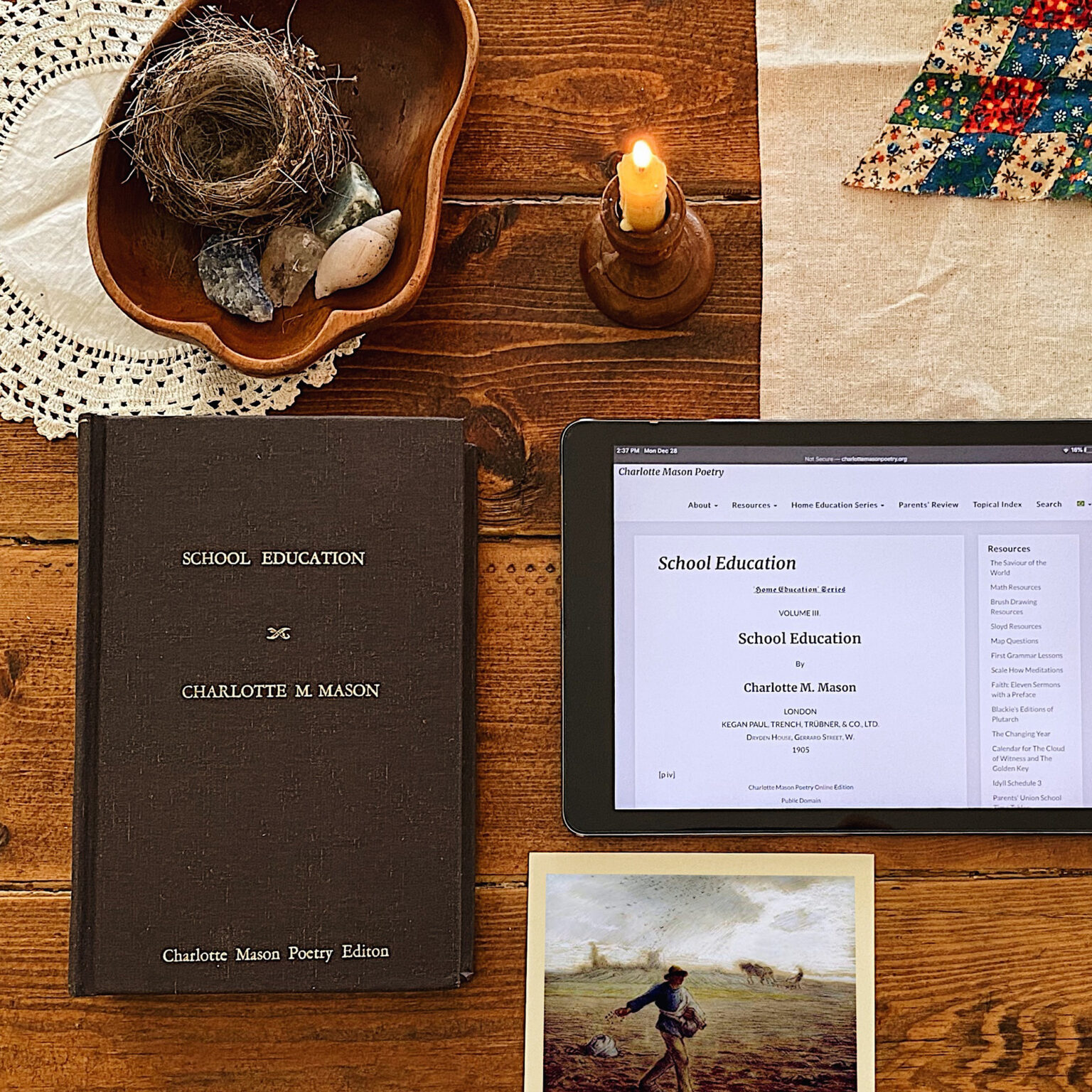 Overhead view of a wooden desk with vintage books, a bird’s nest in a wooden bowl, a lit candle, a quilted corner, and a tablet showing a 'School Education' page.