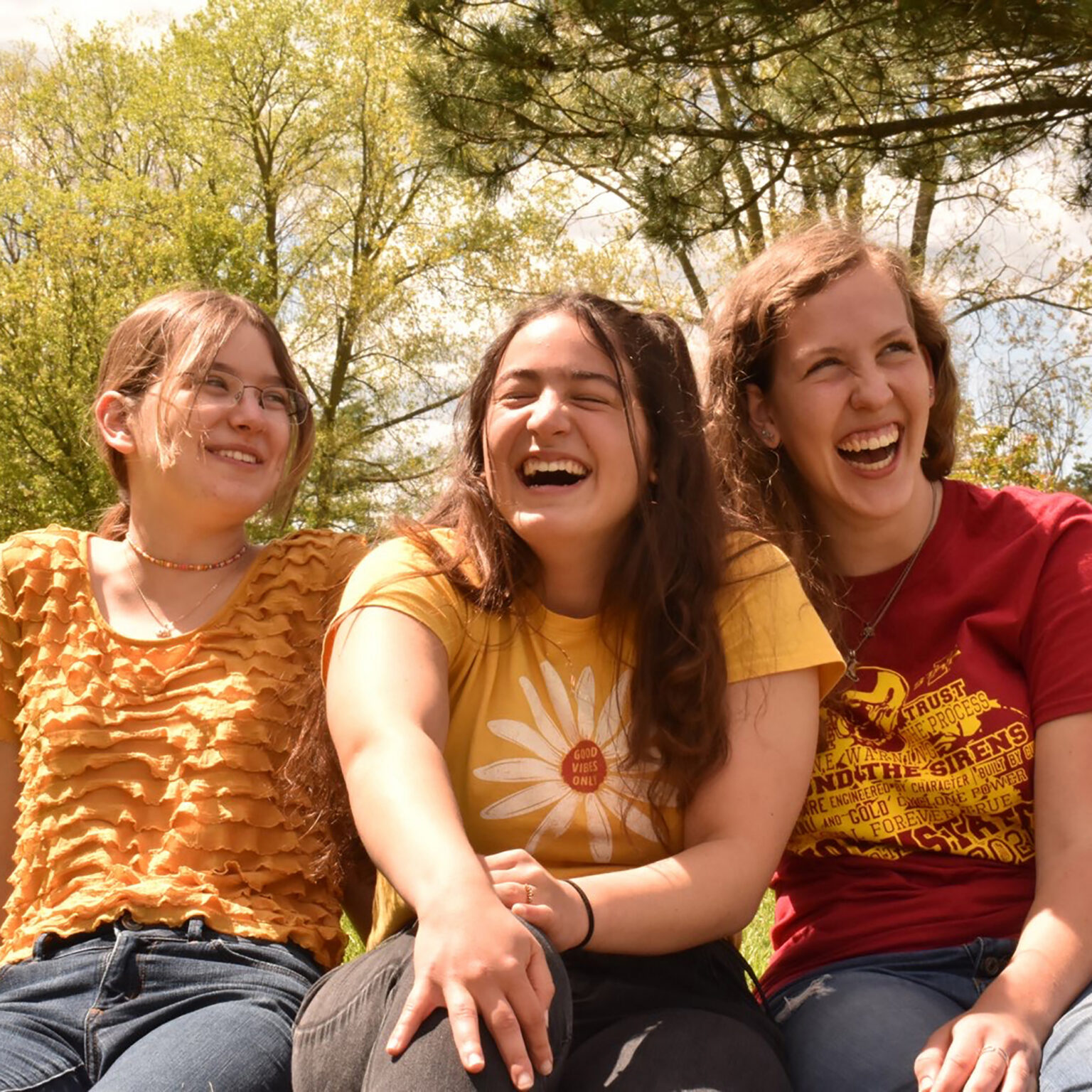 Three young friends sitting outdoors, smiling for a sunny group selfie in casual tops (orange, yellow, red).