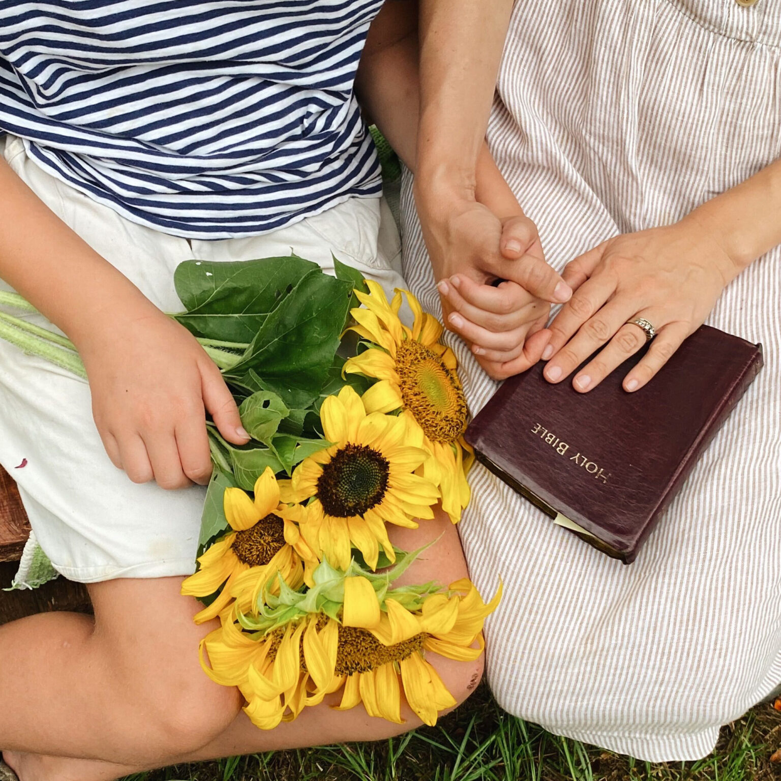 Two people sit side by side, holding hands over a bouquet of sunflowers with a dark red Bible resting nearby.