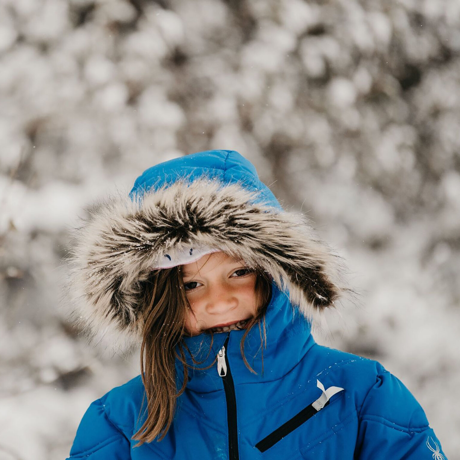 Child in a blue winter jacket with a fur-trimmed hood, smiling in a snowy outdoor scene.