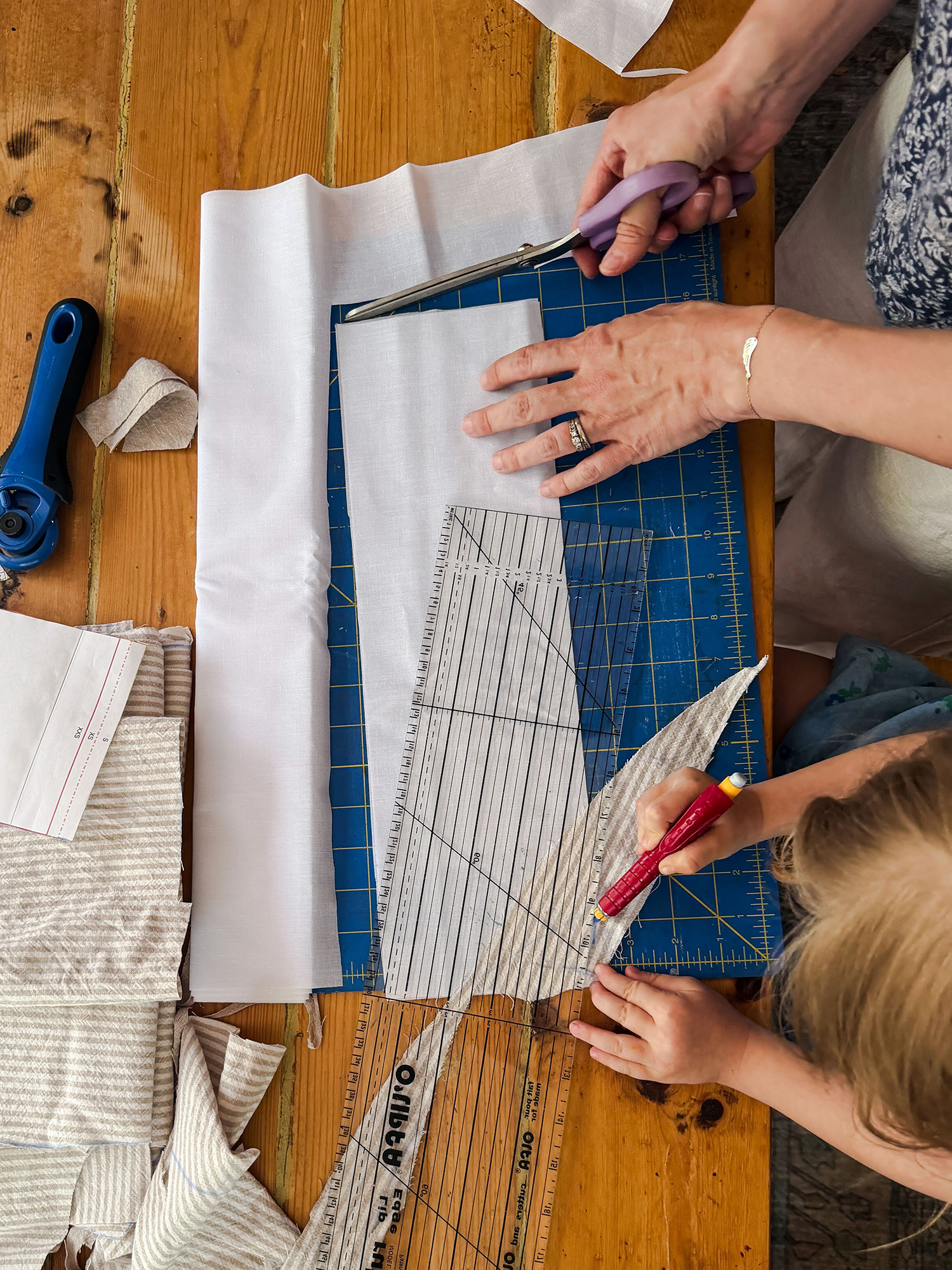 Person cutting light gray fabric with purple-handled scissors on a blue cutting mat, surrounded by rulers and fabric scraps on a wooden table.”](https://)