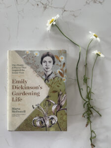 Book cover for Emily Dickinson's Gardening Life placed on a marble surface with white daisies nearby.