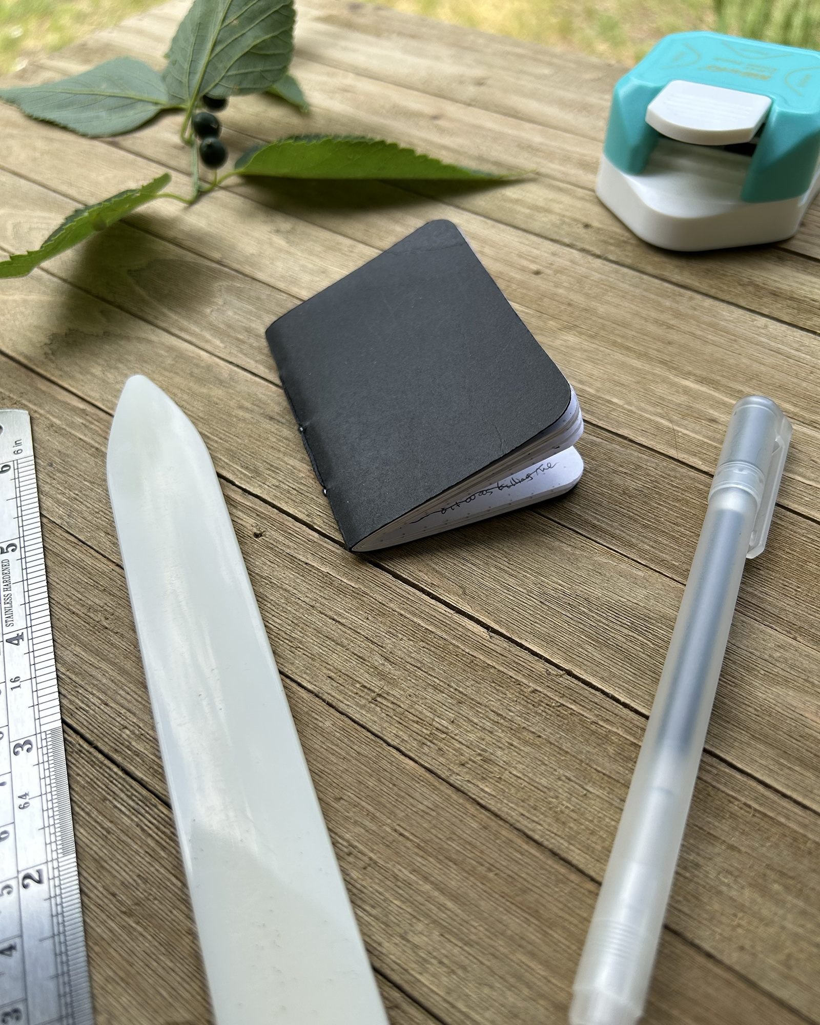 Outdoor wooden table with a small black notebook, ruler, and white/teal tape dispenser alongside green leaves in the background.