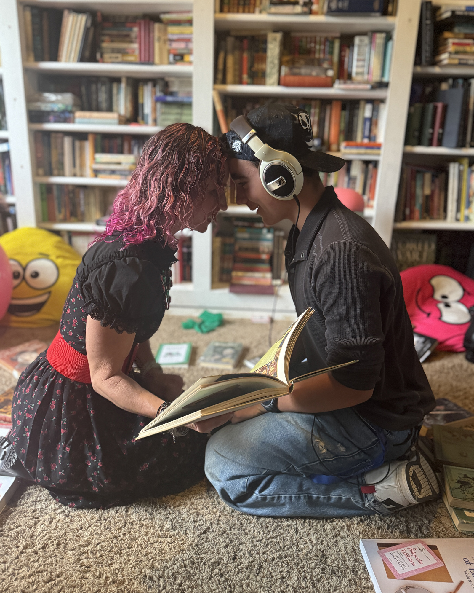 Two people lean their foreheads together over an open book in a cozy library, one with pink-tinted hair and a black dress, the other wearing a cap and headphones.