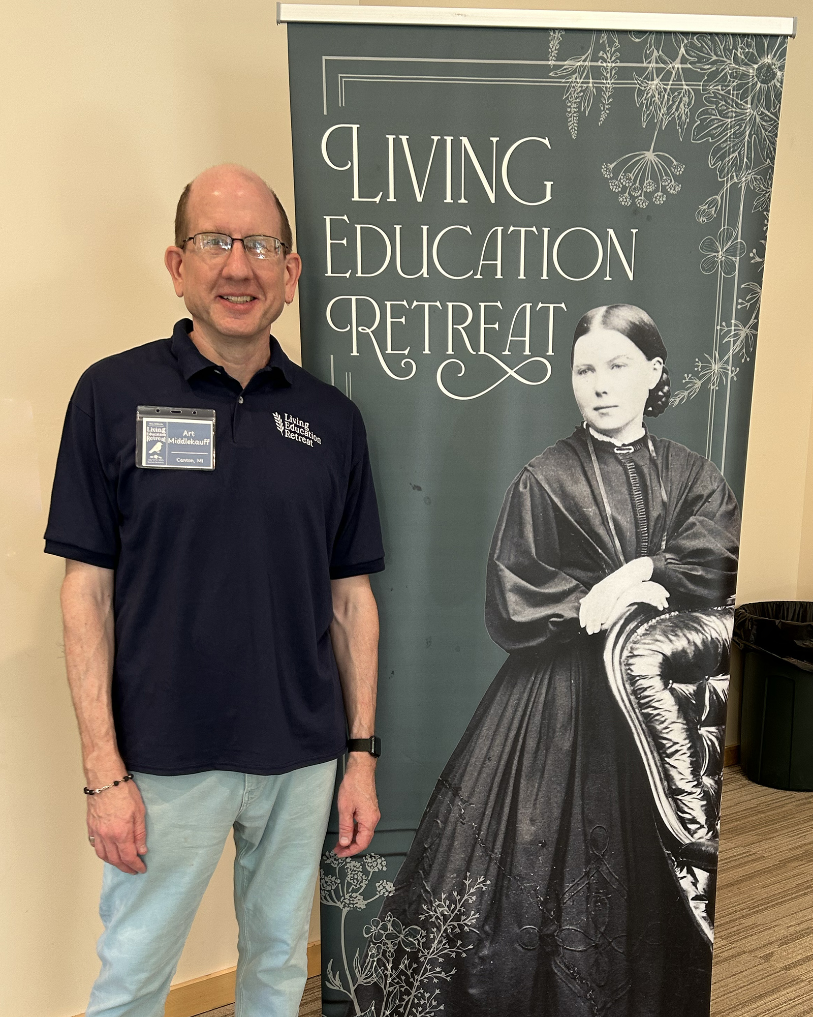 Man in a navy polo and glasses stands beside a tall banner that reads 'Living Education Retreat' with a vintage-style portrait on the right side.
