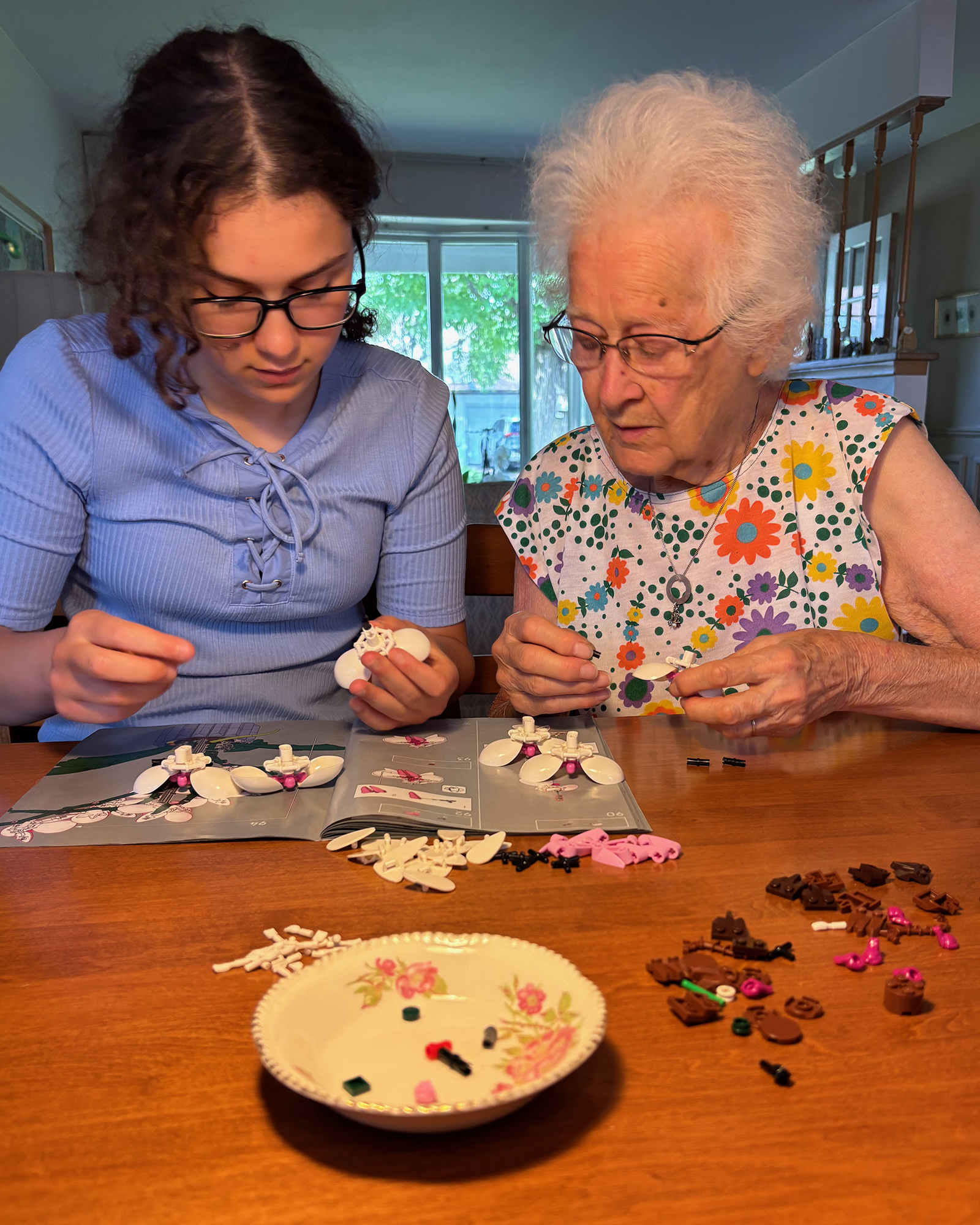 Two women, a younger girl and an elderly woman, concentrating while assembling small plastic model pieces at a table indoors.