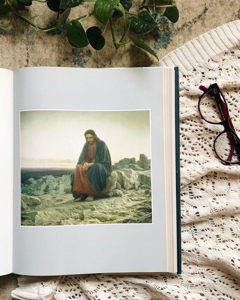 Open book showing a painting of a bearded man sitting on rocks by the sea, with a plant and lace fabric in the background and glasses to the right.