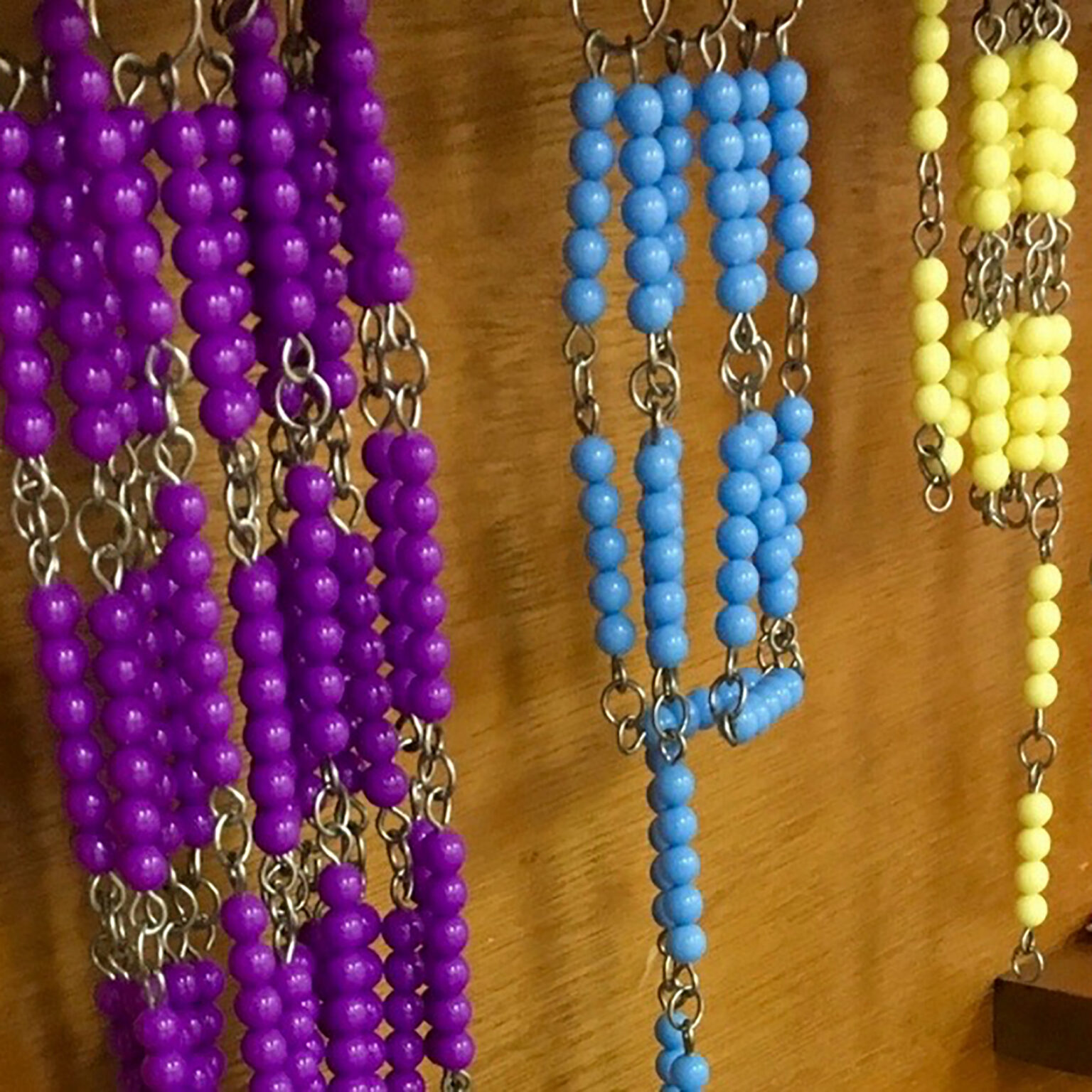Rows of colorful bead necklaces (purple, blue, yellow) hanging on hooks against a wooden backdrop.