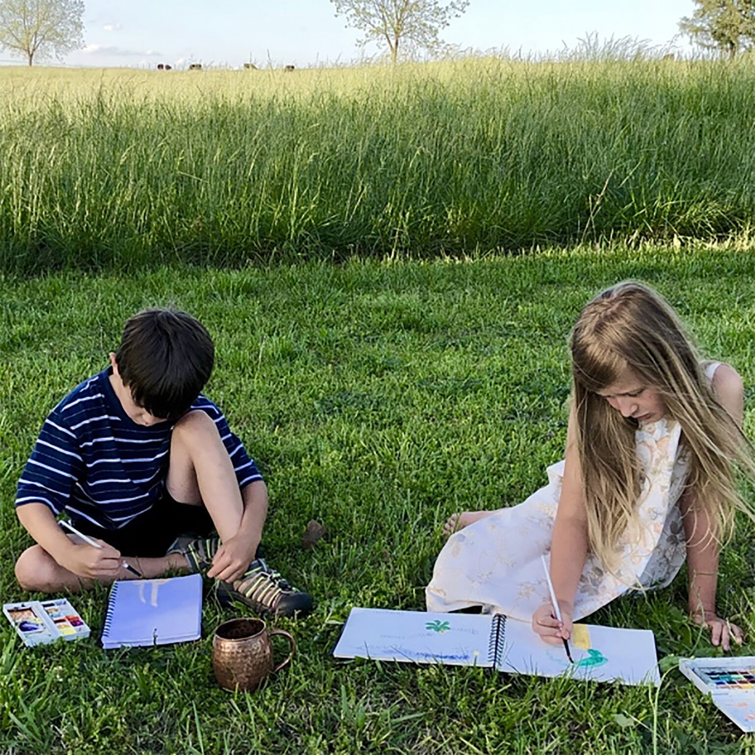 Two children drawing on the grass in a park, with open sketchbooks, paints, and brushes nearby under a sunny sky.