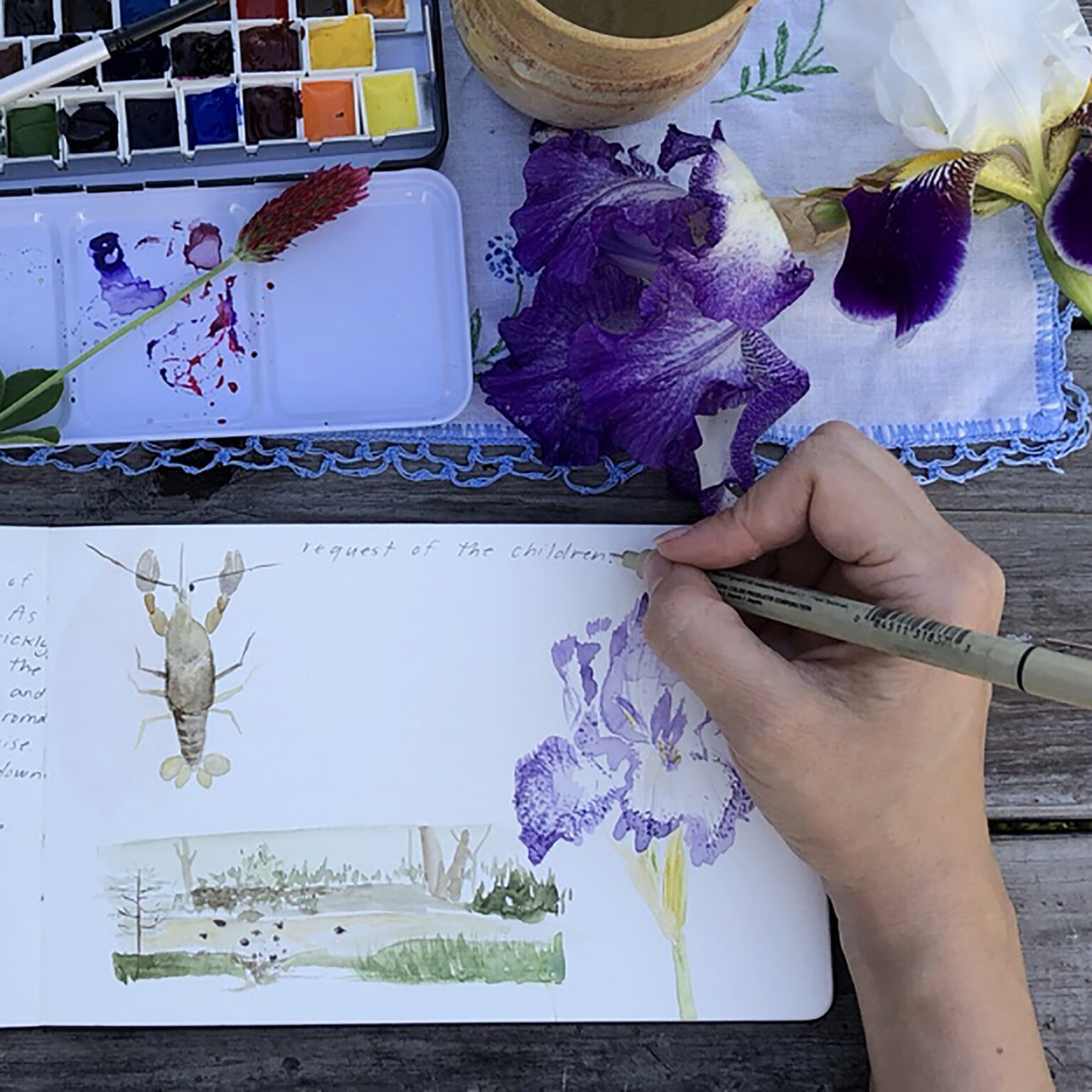 Watercolor paints and brush with a nearby cup, purple irises on a floral tablecloth, and a hand drawing a plant in a sketchbook nearby.