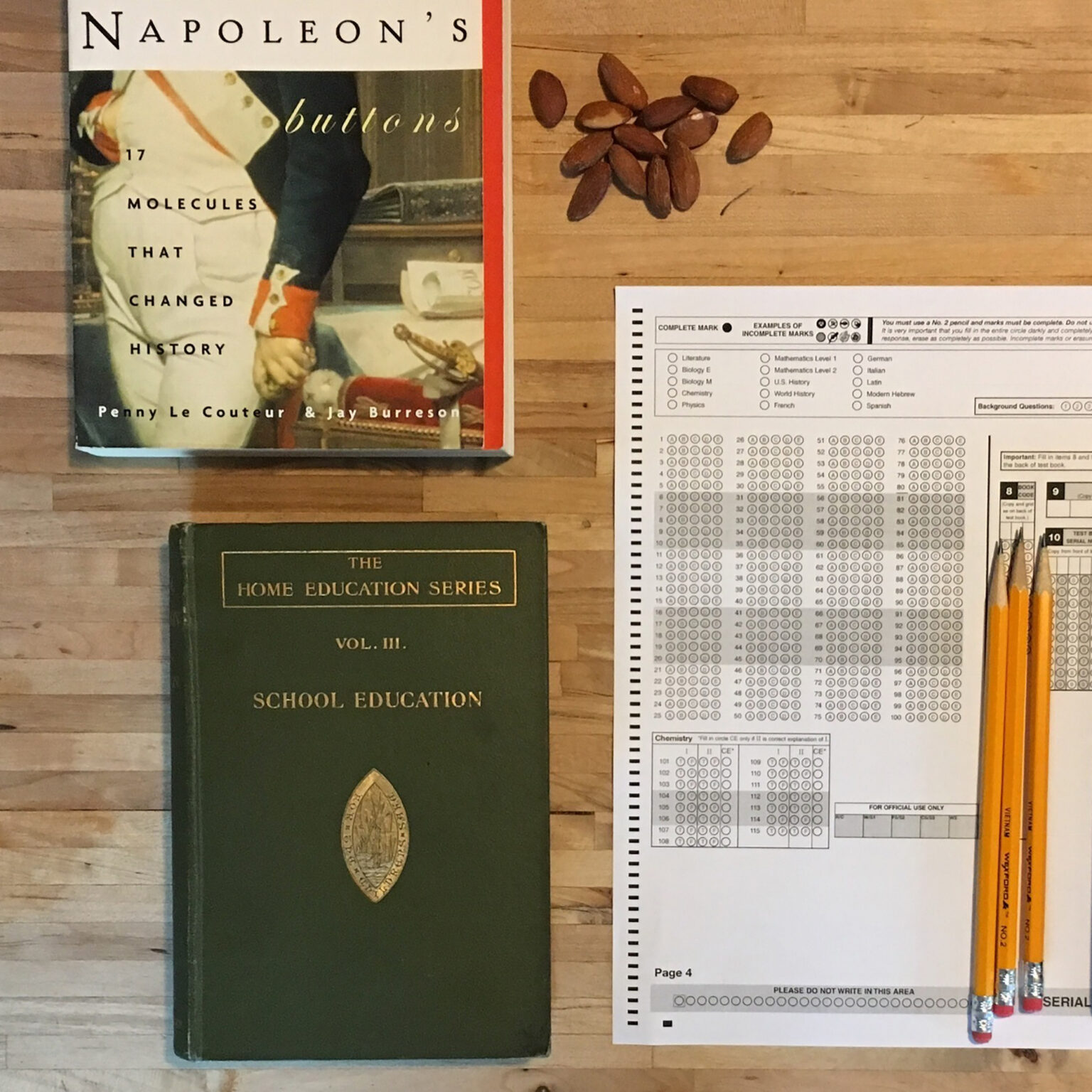 Book covers on a wooden desk: 'Napoleon's Buttons' and 'Molecules That Changed History' beside a dark green 'Home Education Series' volume, with a test sheet, pencils, and a cluster of almonds nearby.