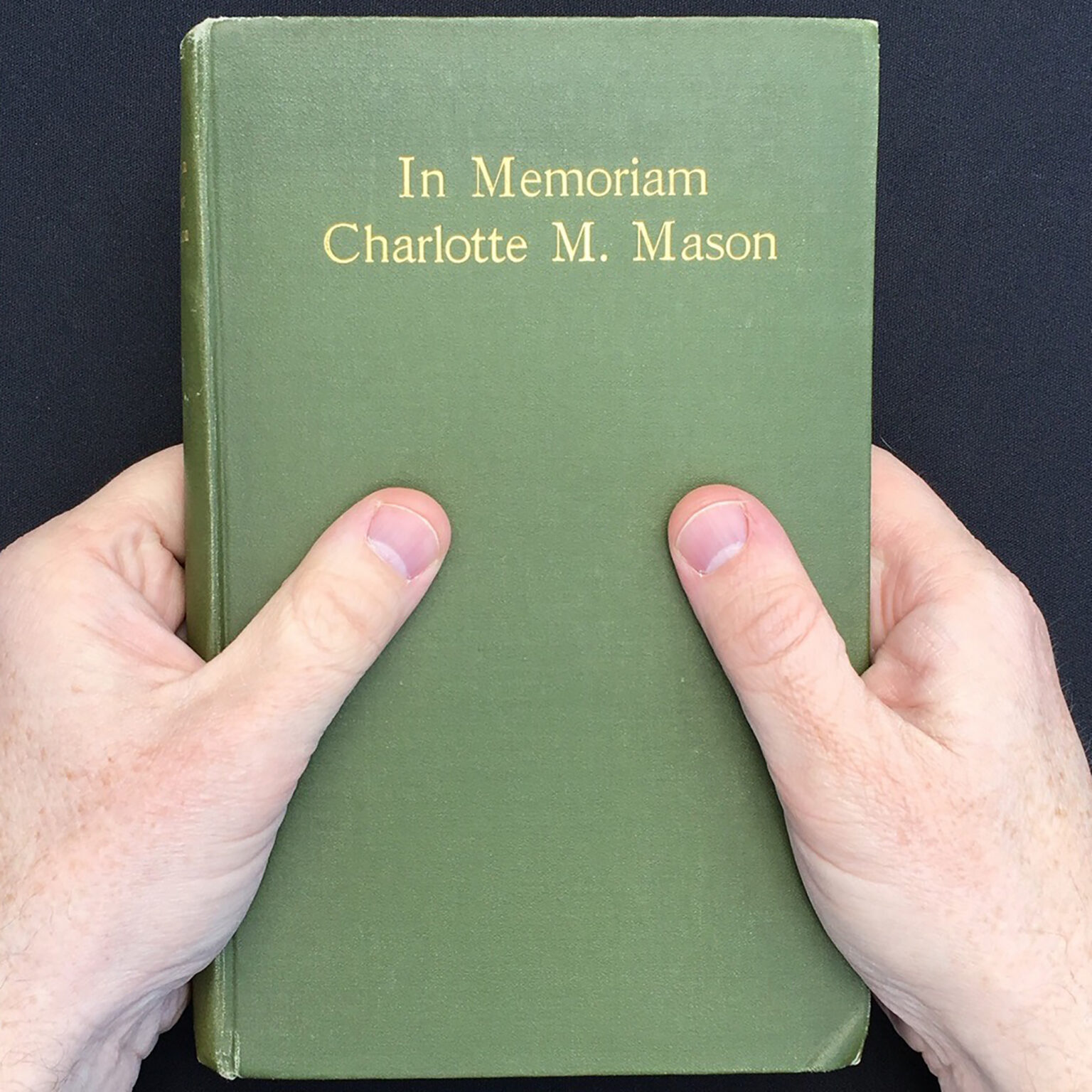 Green hardcover book held by both hands against a dark fabric background, bottom corners visible.