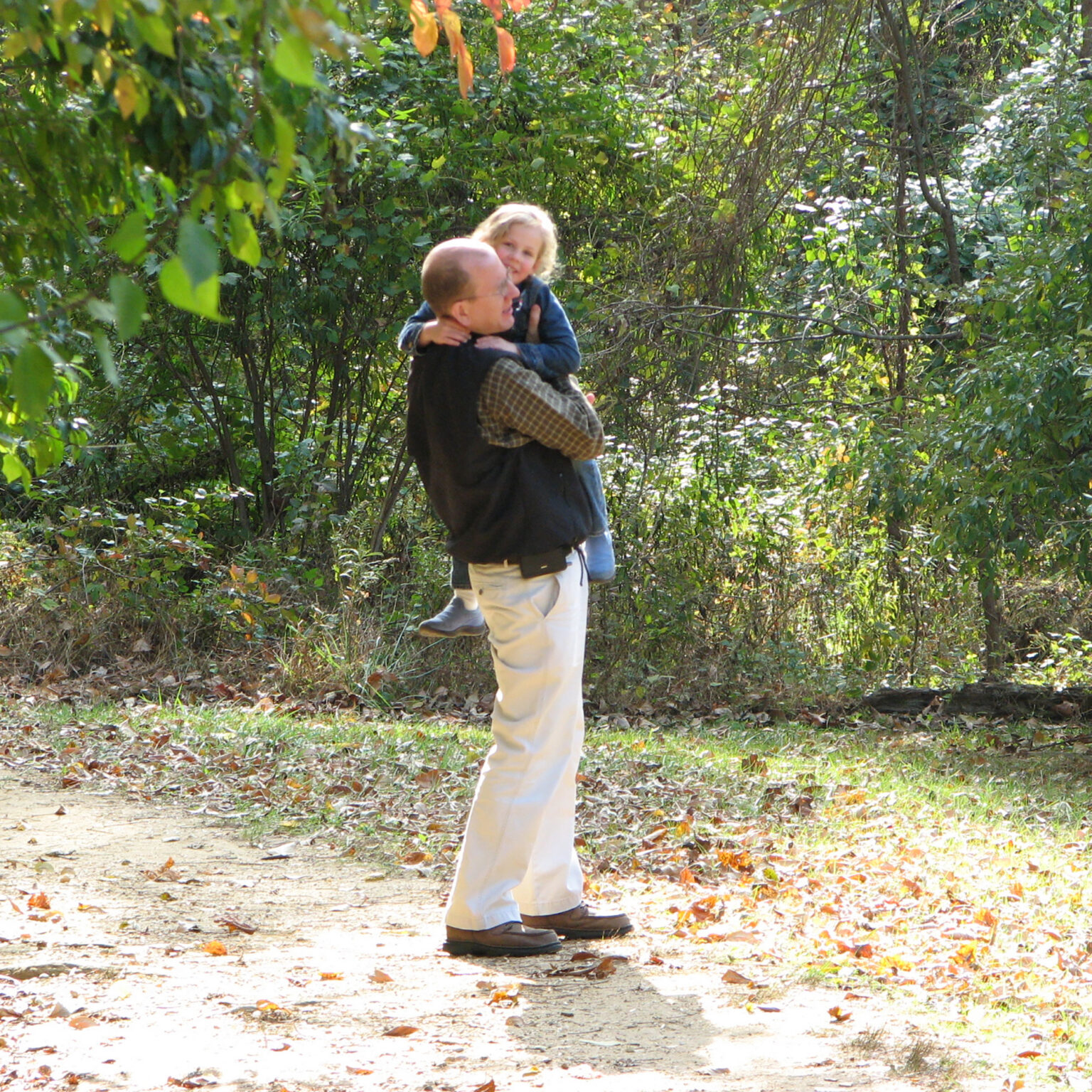 Adult man lifting a smiling child in a dense, leafy area with autumn leaves on the ground.