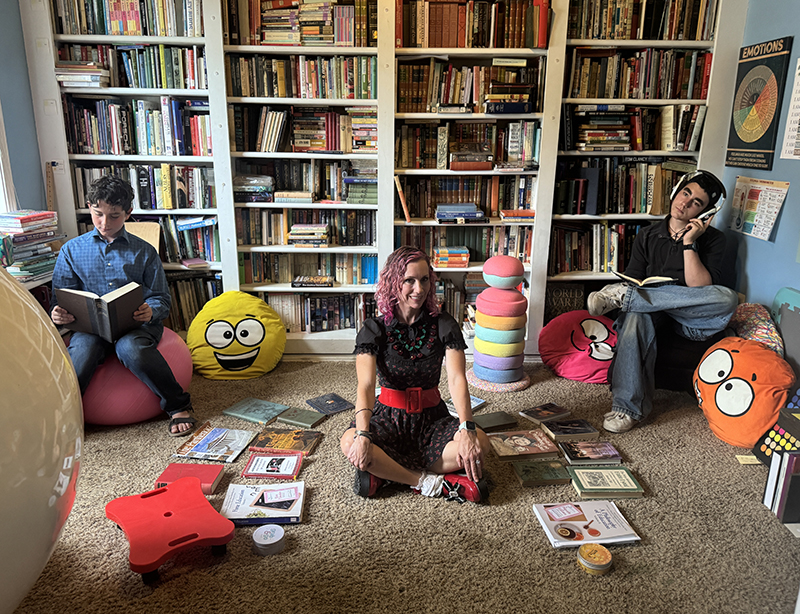 Three people sit among floor books in a cozy library: a woman in the center on the carpet, flanked by two readers on cushions and a wall of shelves behind.