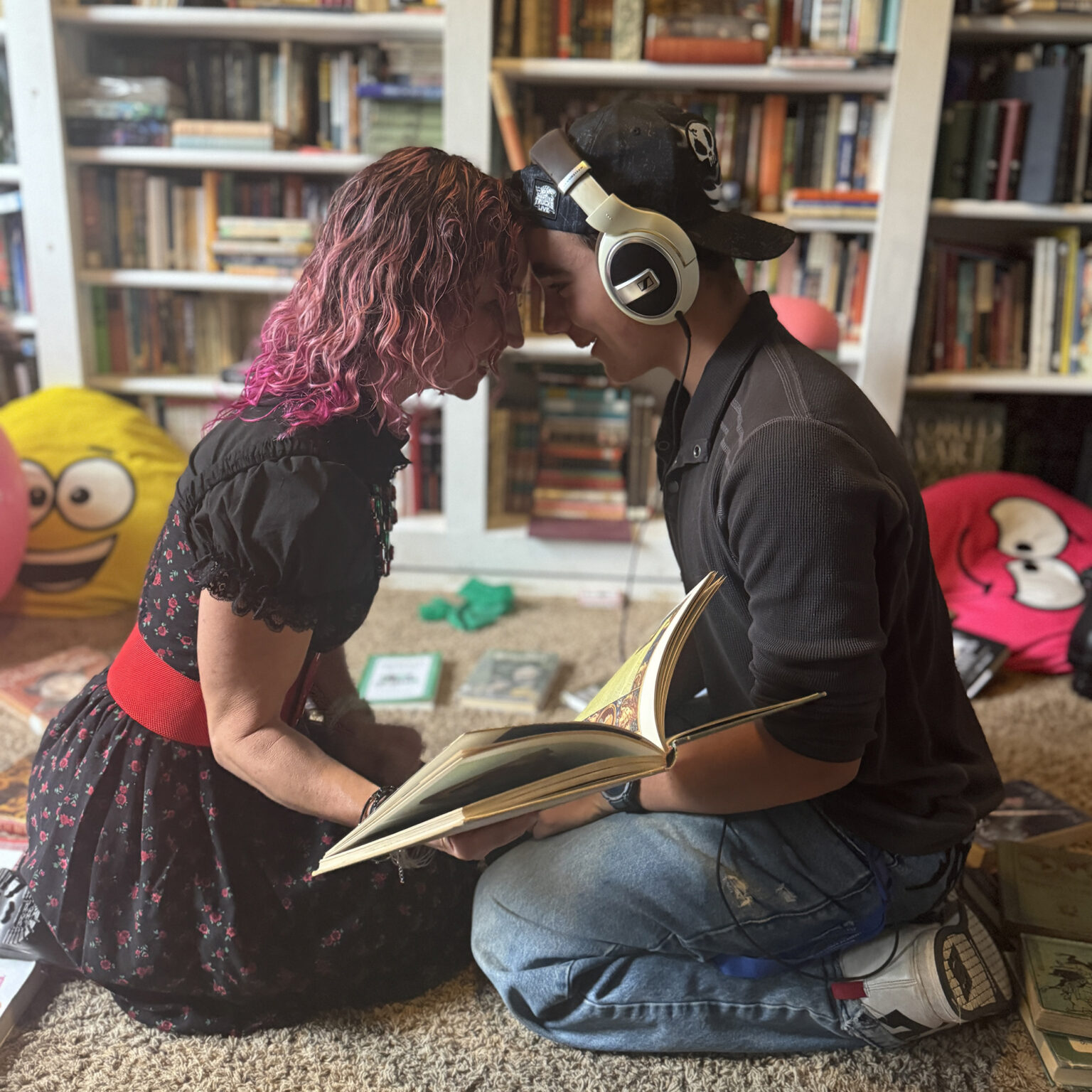 Two people lean in with foreheads touching, wearing large over-ear headphones, looking at an open book in a cozy, book-filled room.