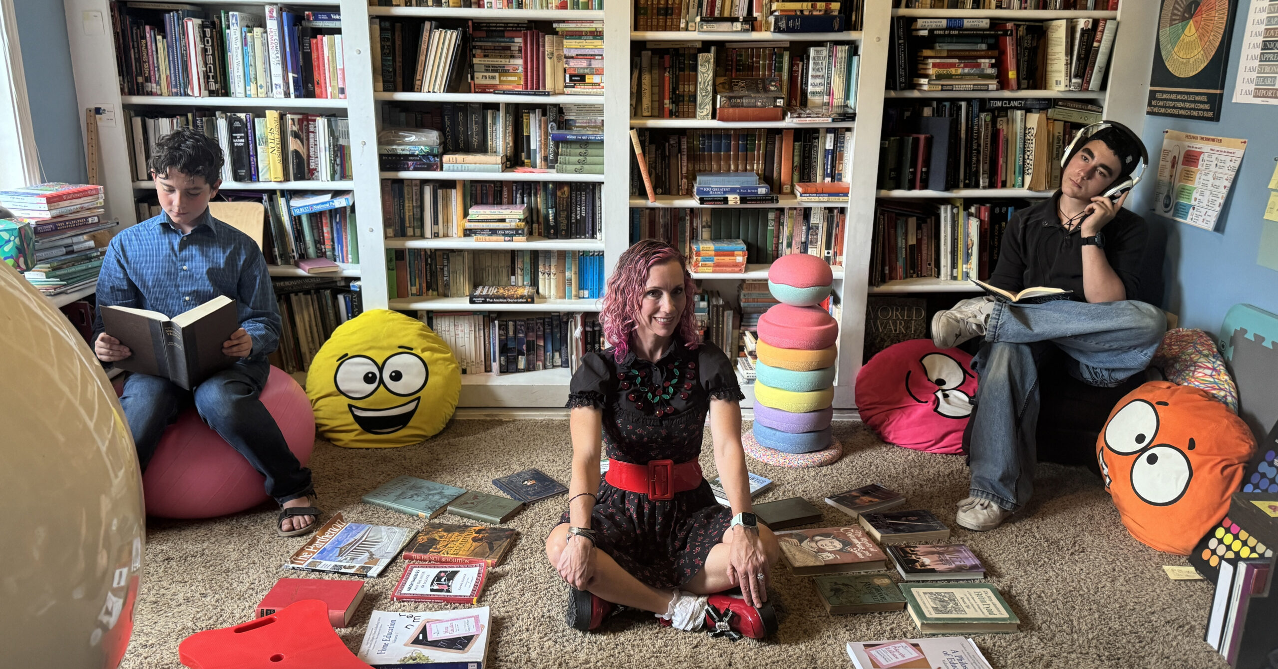Three people sit among floor cushions and a wall of books; a boy on the left reads a book, a woman with pink hair sits center, and a person with headphones reads on the right.