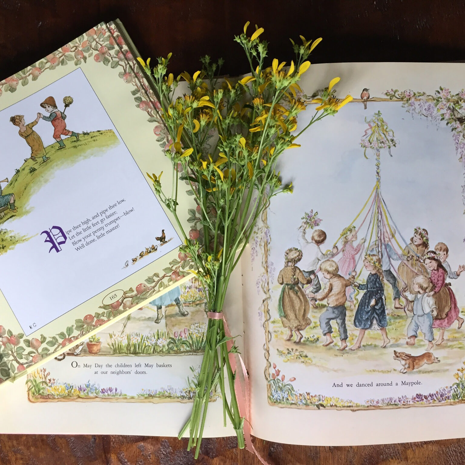 Open vintage children's book spread showing children dancing with floral borders; a yellow bouquet rests on the pages as if laid there for display.