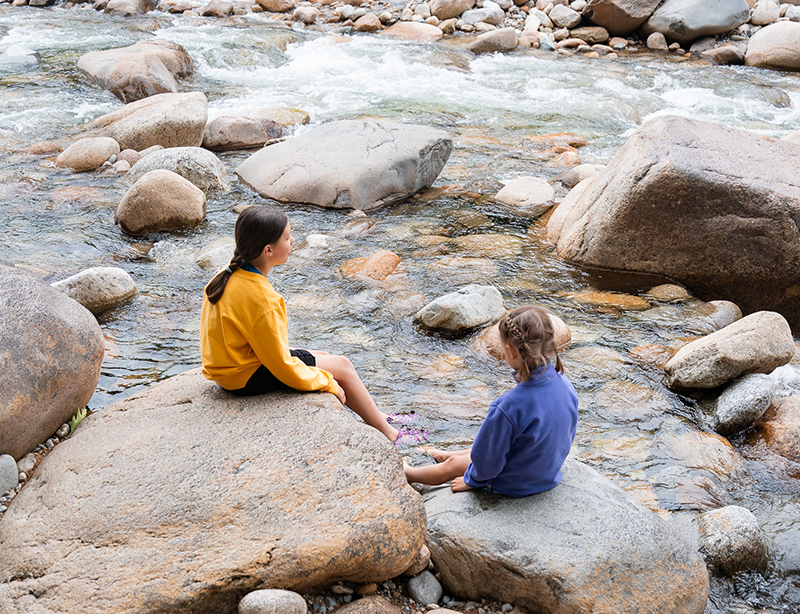 Two children sit on large rocks beside a flowing river, one in a yellow sweater and the other in blue.