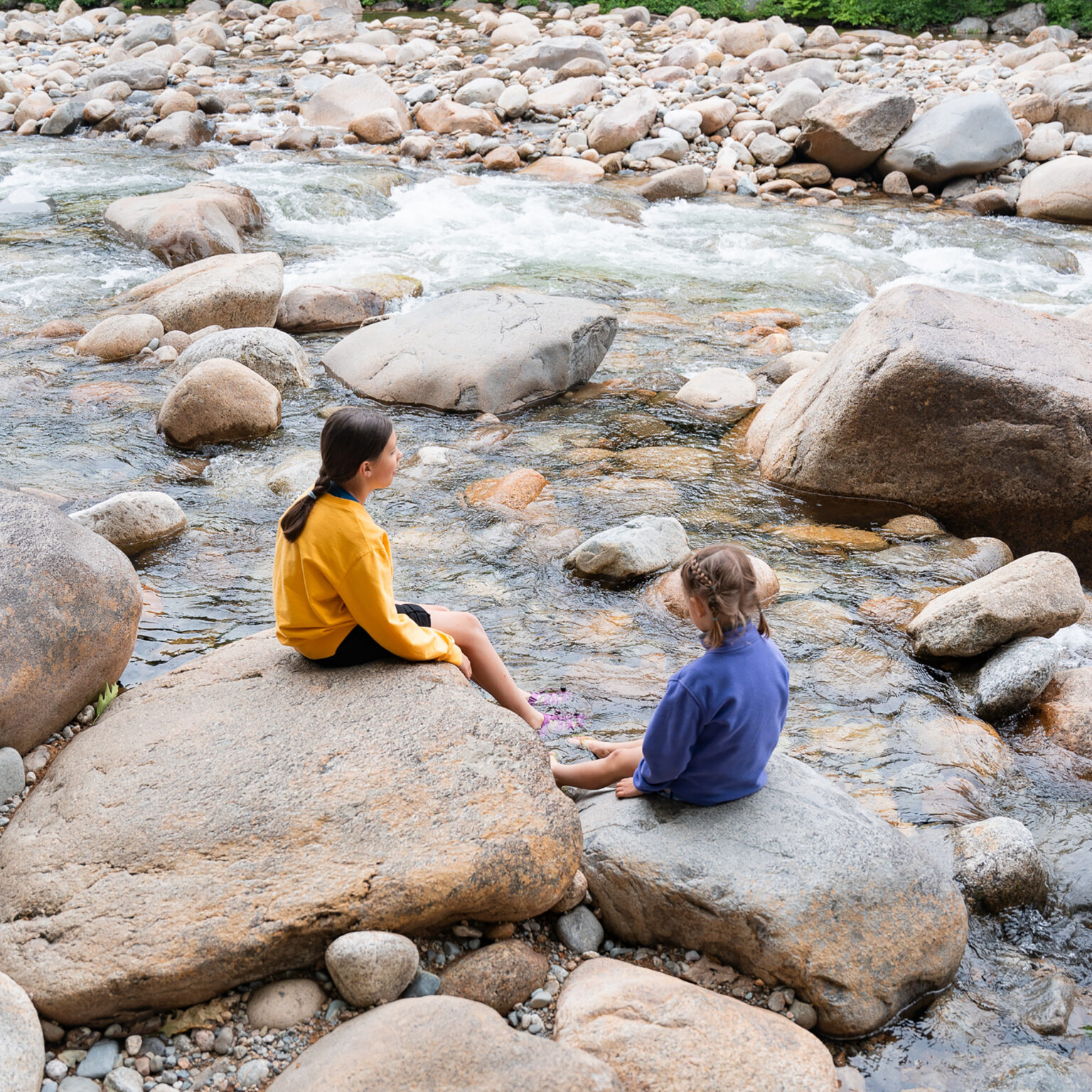 Two children sit on large rocks beside a fast-flowing rocky river, surrounded by smooth, rounded stones and pebbles.