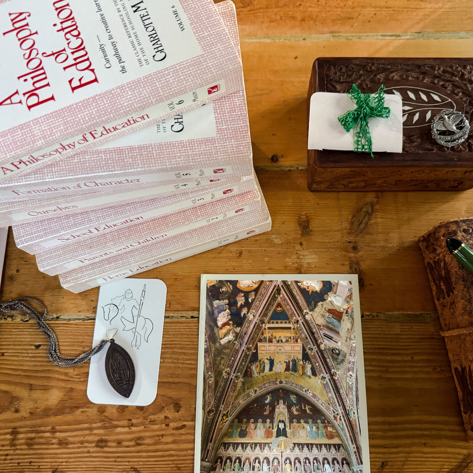 Stack of pink-patterned education textbooks on a wooden table, with a white gift box tied with a green ribbon and a metal medallion to the right. A decorative book page shows ornate artwork nearby.