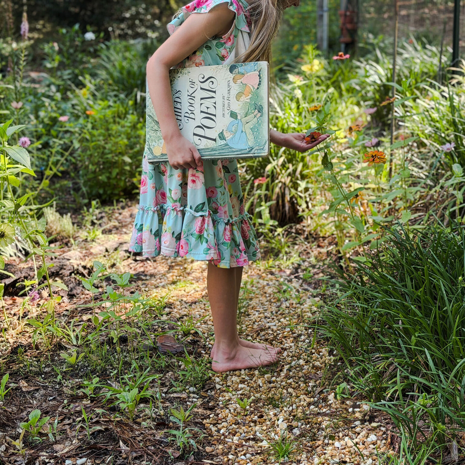 Girl in a floral dress stands barefoot in a garden, holding a large illustrated book titled 'Poems' under her arm while picking a flower with her other hand.