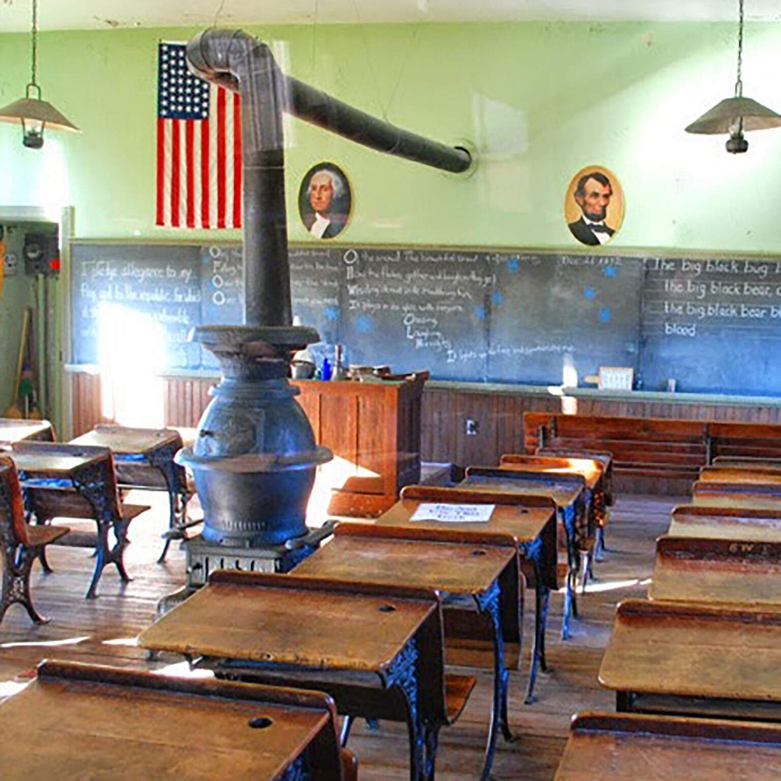 Old-fashioned classroom with wooden desks, a large stove, and an American flag on the wall.