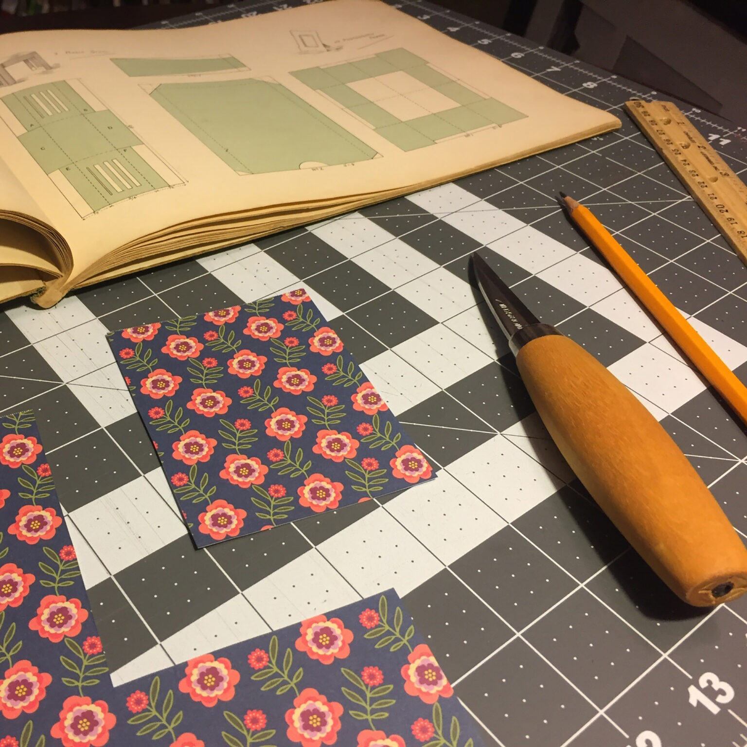 Open design book on a cutting mat showing green layout diagrams, with a ruler and pencil nearby.