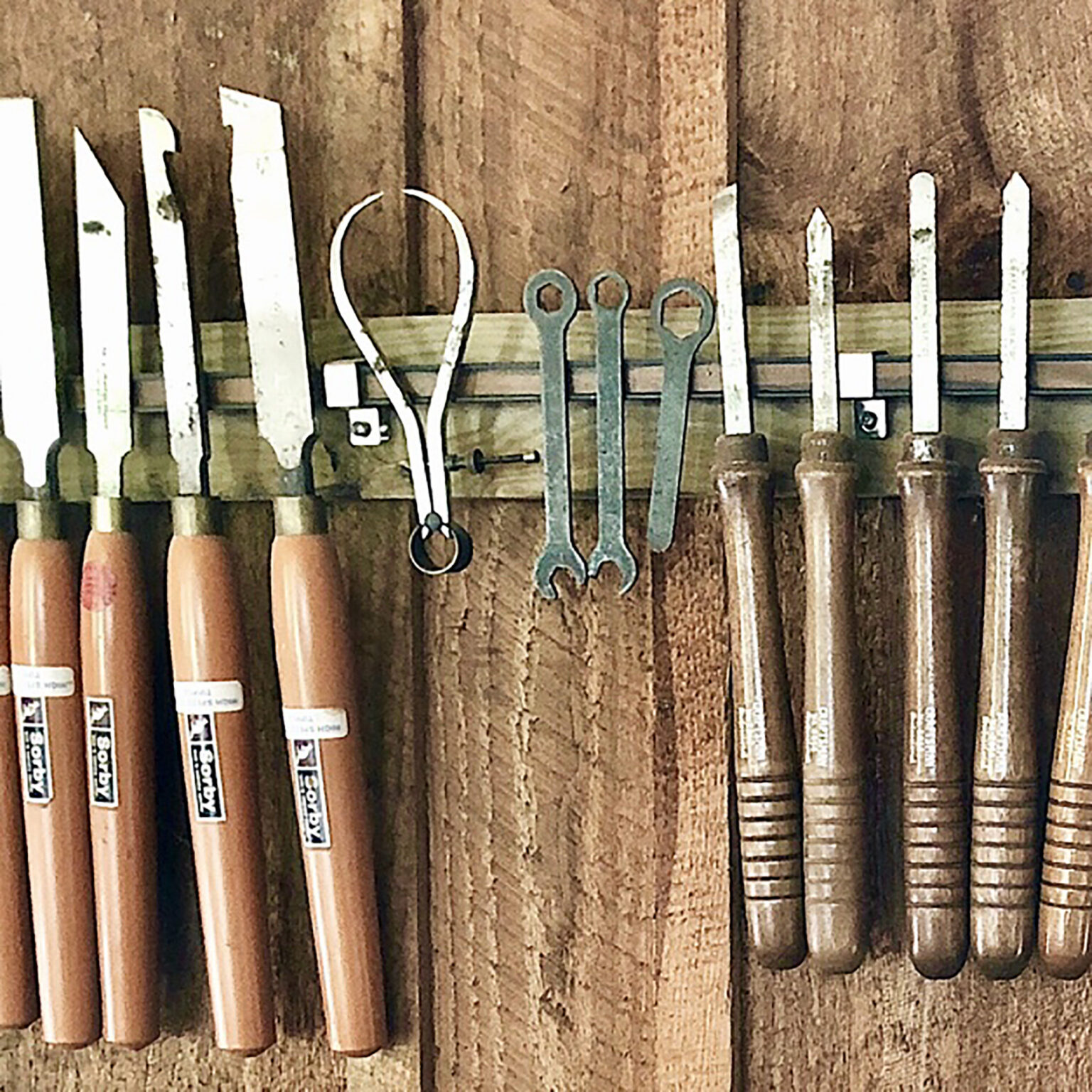 Row of hand tools hanging on a wooden board, including scissors, wrenches, and chisels in a workshop setting.