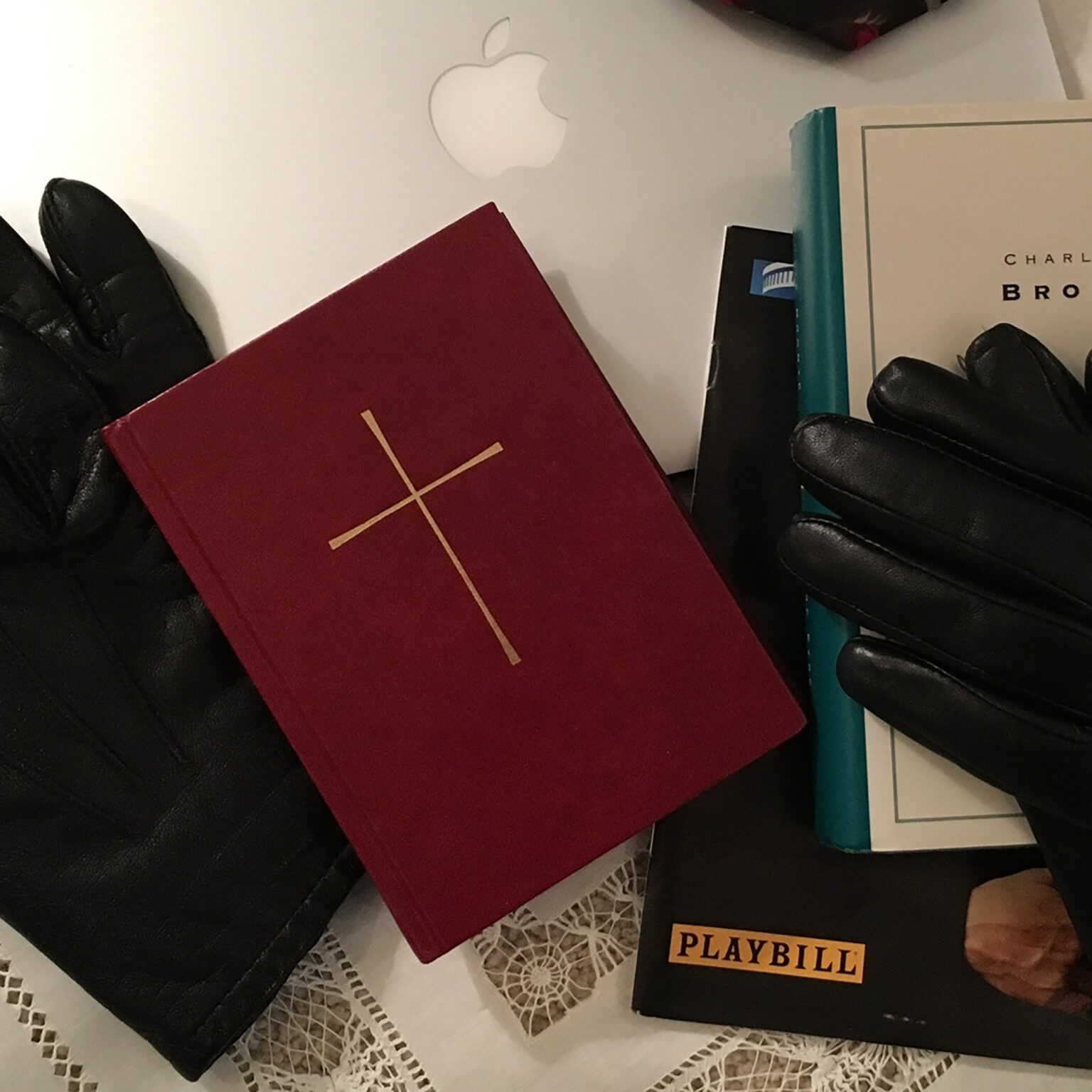 Top-down view of assorted items on a table: black gloves, a burgundy notebook, a turquoise binder, a Playbill booklet, and a white lace tablecloth with an Apple logo in the background
