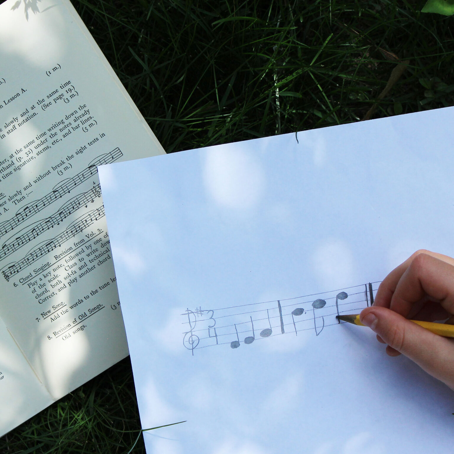 Outdoor scene with papers: music manuscript and printed text on grass, a hand holding a yellow pencil nearby