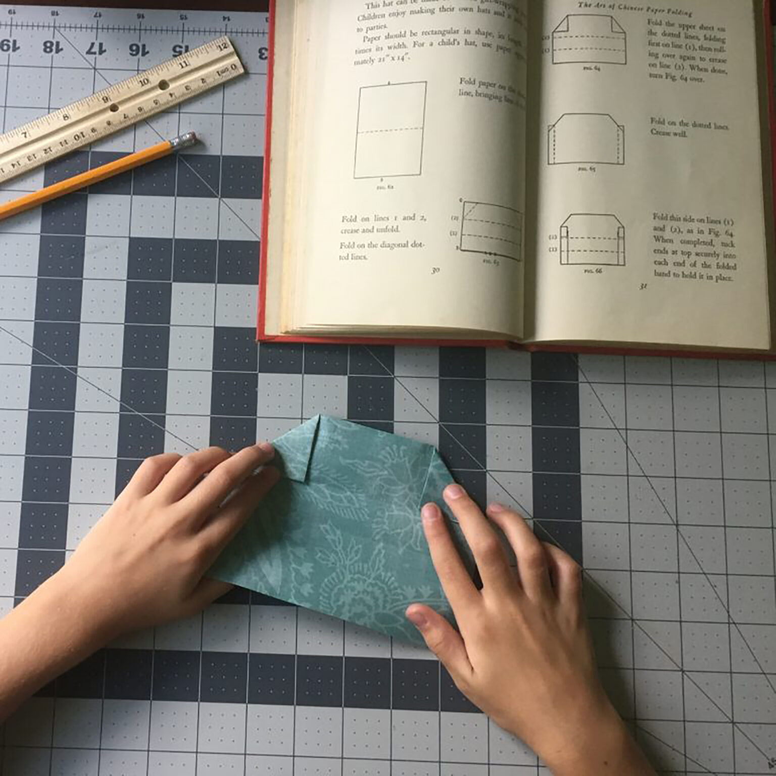 Person folds a turquoise origami square on a cutting mat beside an open craft book with folding diagrams; a ruler and pencil lie nearby.