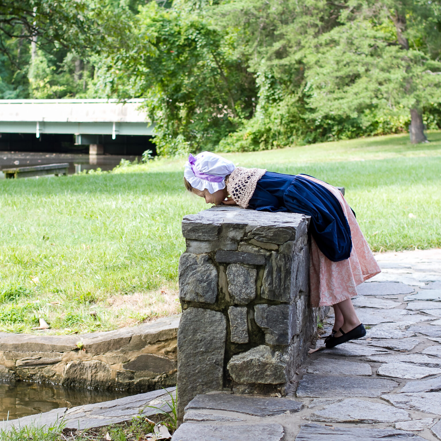 Person in a vintage dress and bonnet leaning over a stone wall by a grassy park and calm water in the background