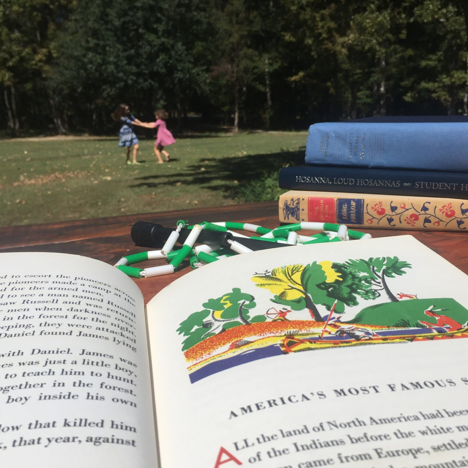 Open book in foreground with an illustrated page; two children in dresses play and dance on a grassy lawn in the background, while a stack of books sits on a table nearby.