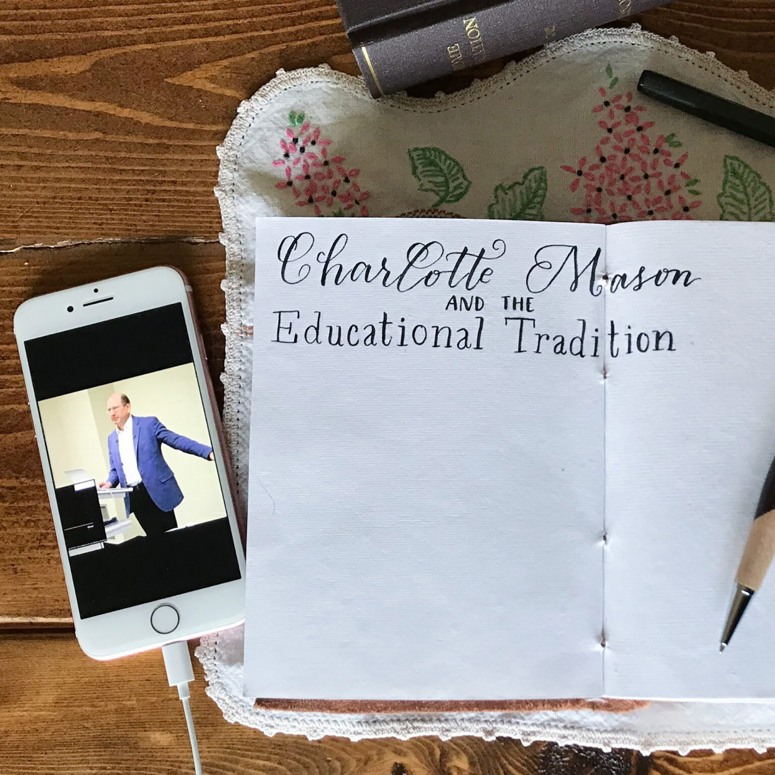Smartphone showing a man in a blue suit giving a talk, placed on a notebook with 'Charlotte Mason' title, on a wooden table.