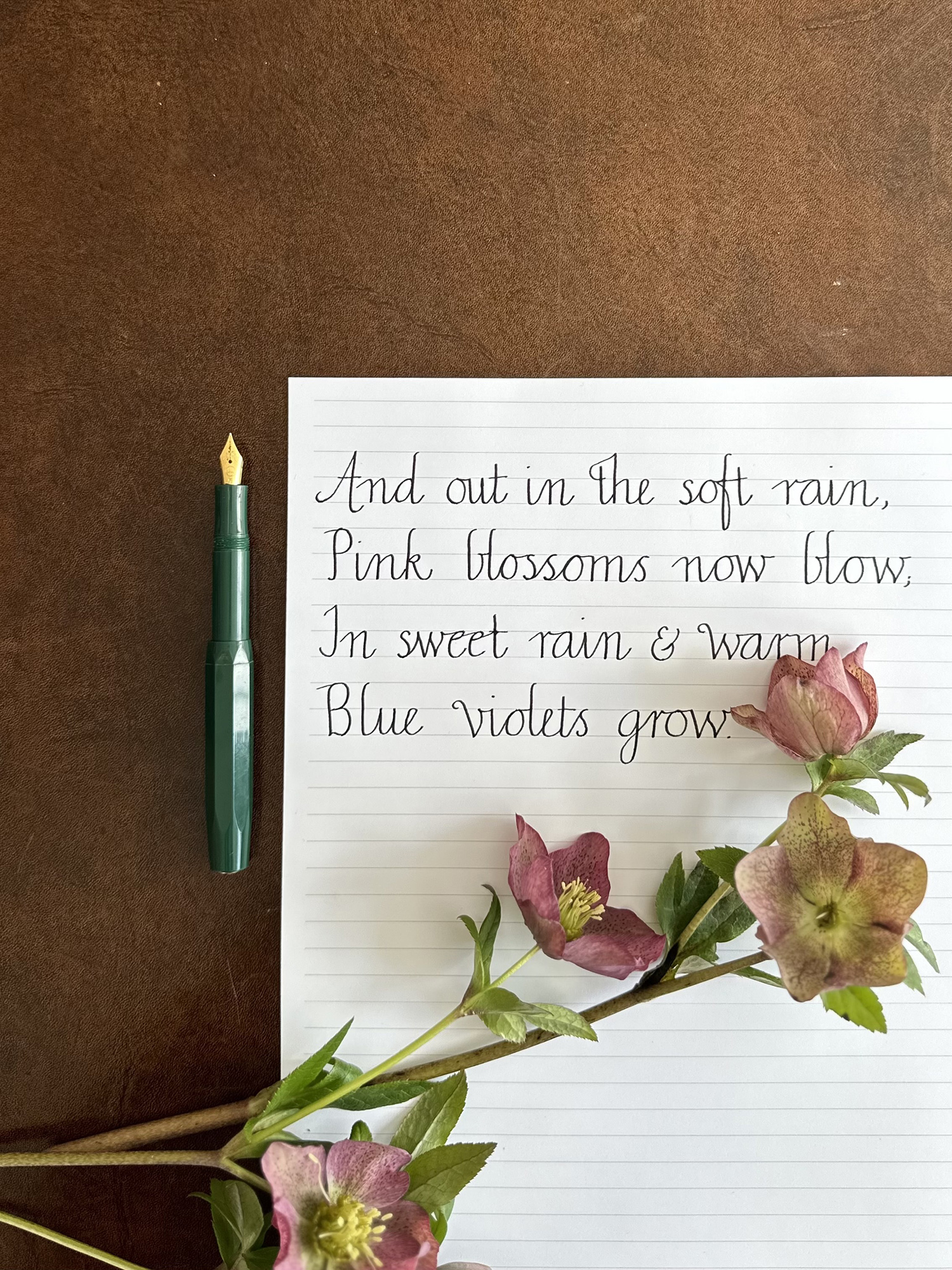 Green fountain pen beside a lined notebook with handwritten cursive text and pink flowers on a brown desk surface.