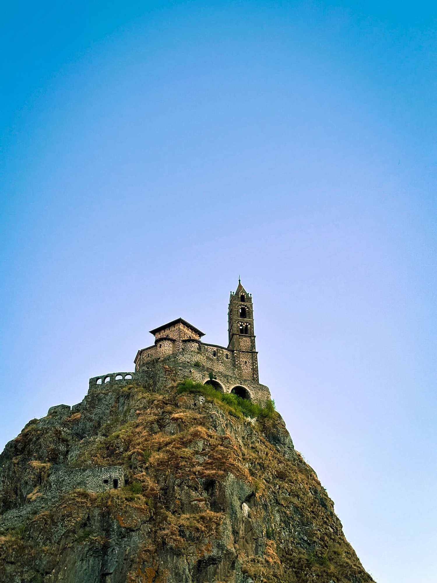 Stone hilltop fortress with a tall tower perched on a rocky cliff against a clear blue sky.