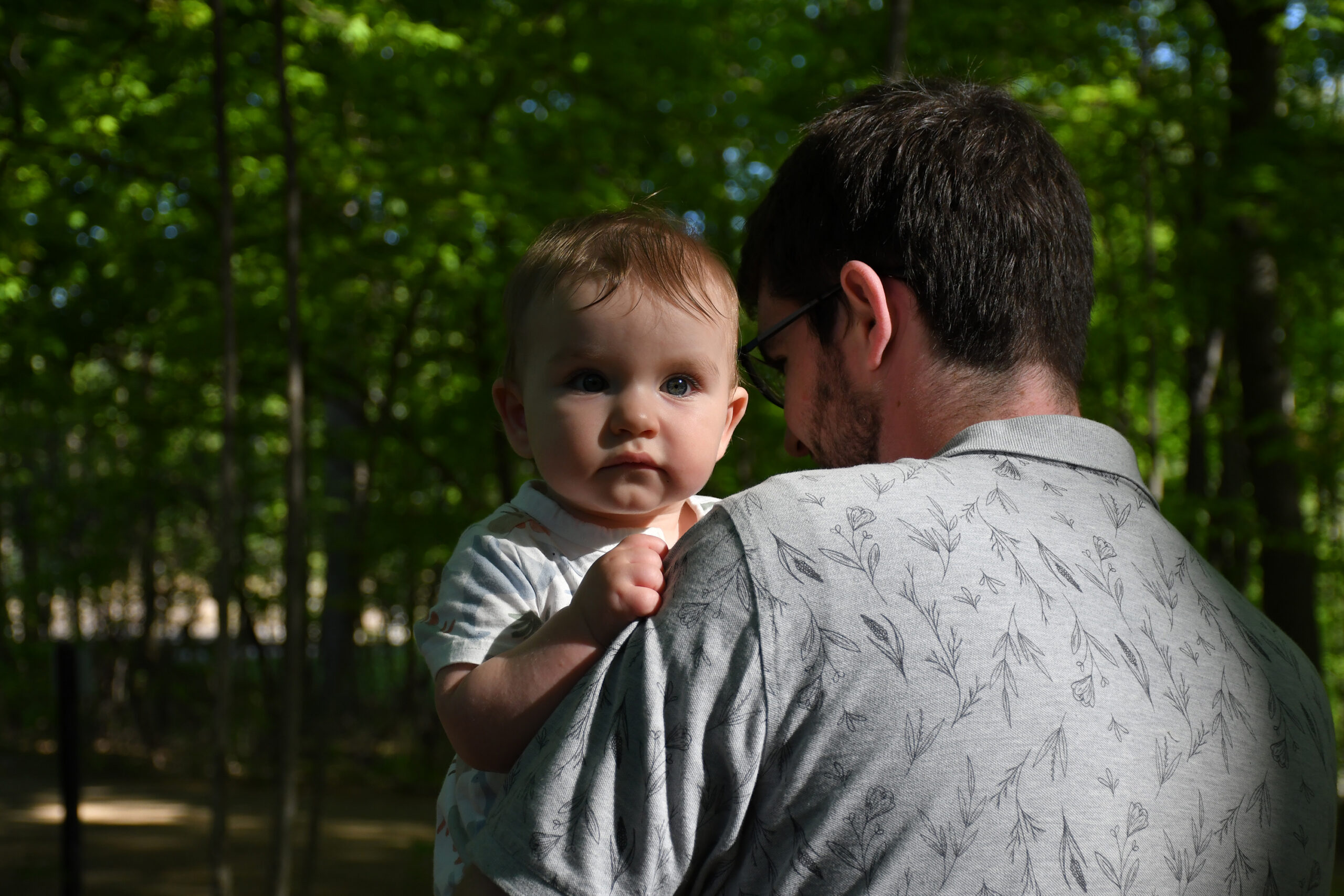 Adult man wearing a patterned shirt holds a baby in a forest, baby looking toward the camera as sunlight filters through trees