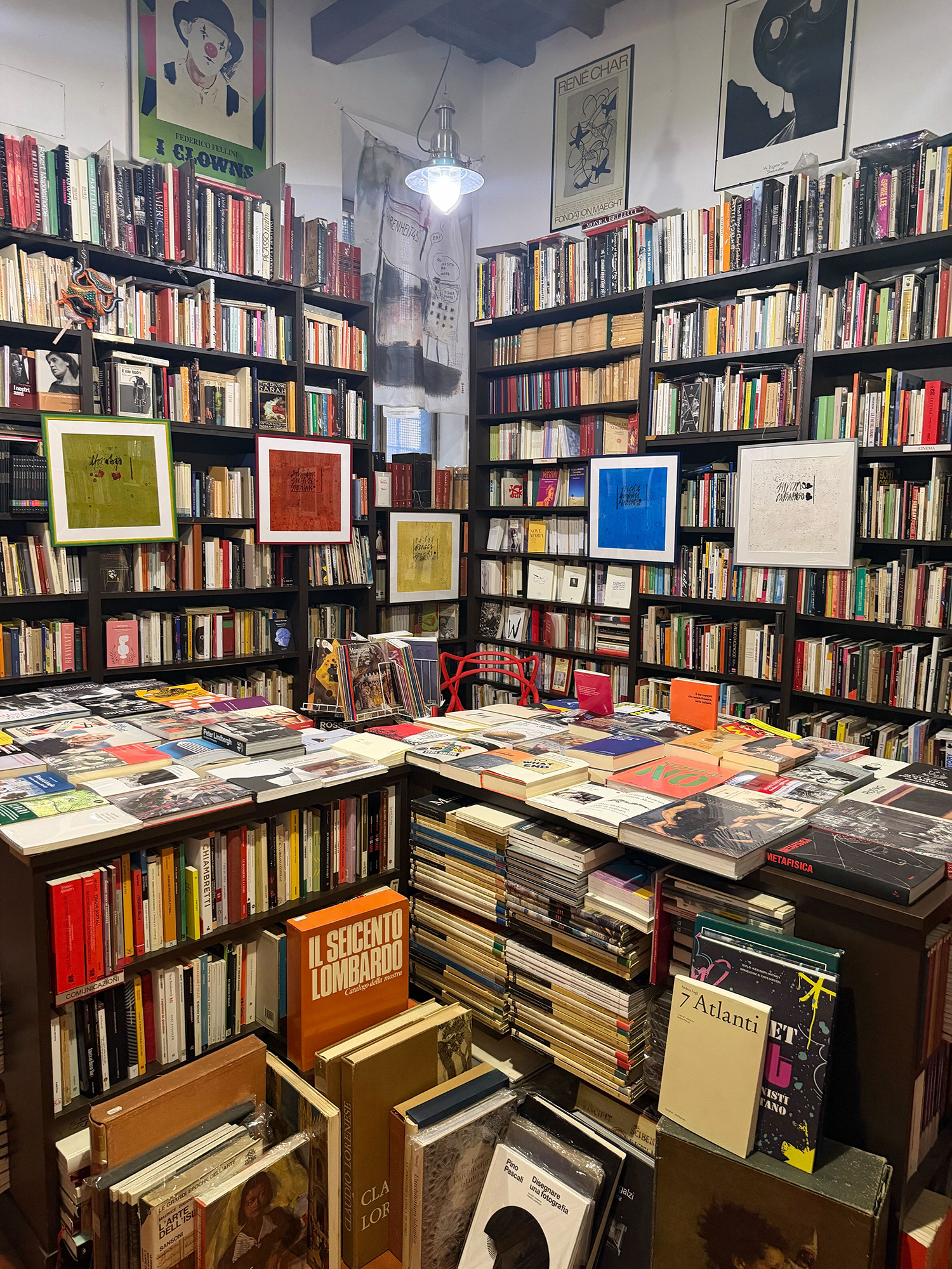 Interior of a crowded bookstore with tall shelves of books and colorful framed art on display tables in the foreground