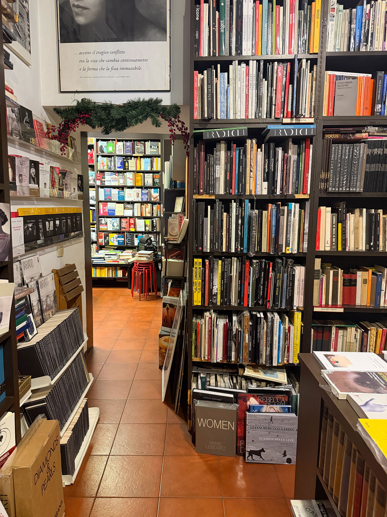 Narrow bookstore aisle with tall shelves of books on the right, warm terracotta floor, and a garlanded doorway leading to a back room.