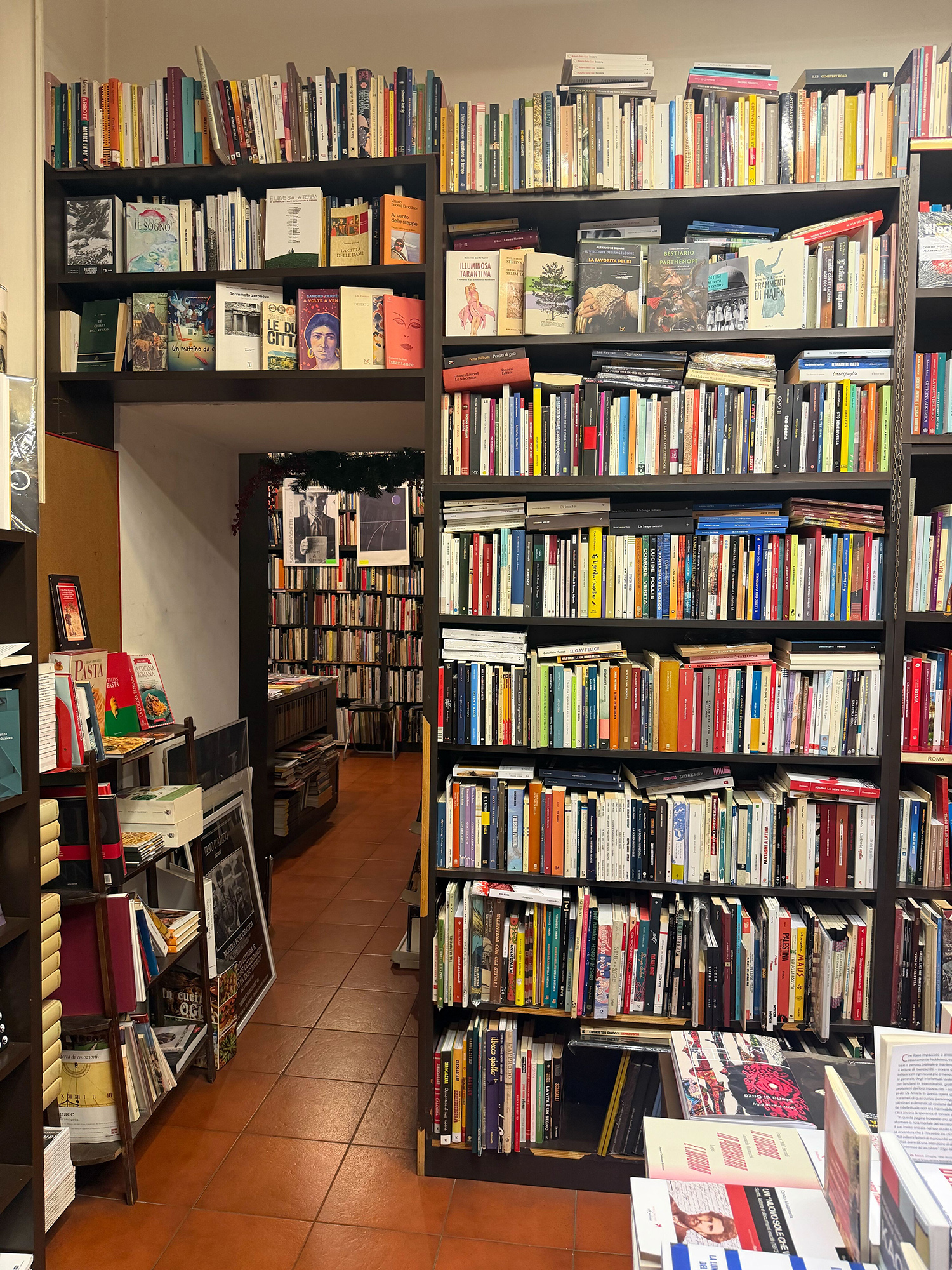 Narrow bookstore aisle with tall black shelves filled with colorful books on both sides, orange tile floor visible in the foreground.