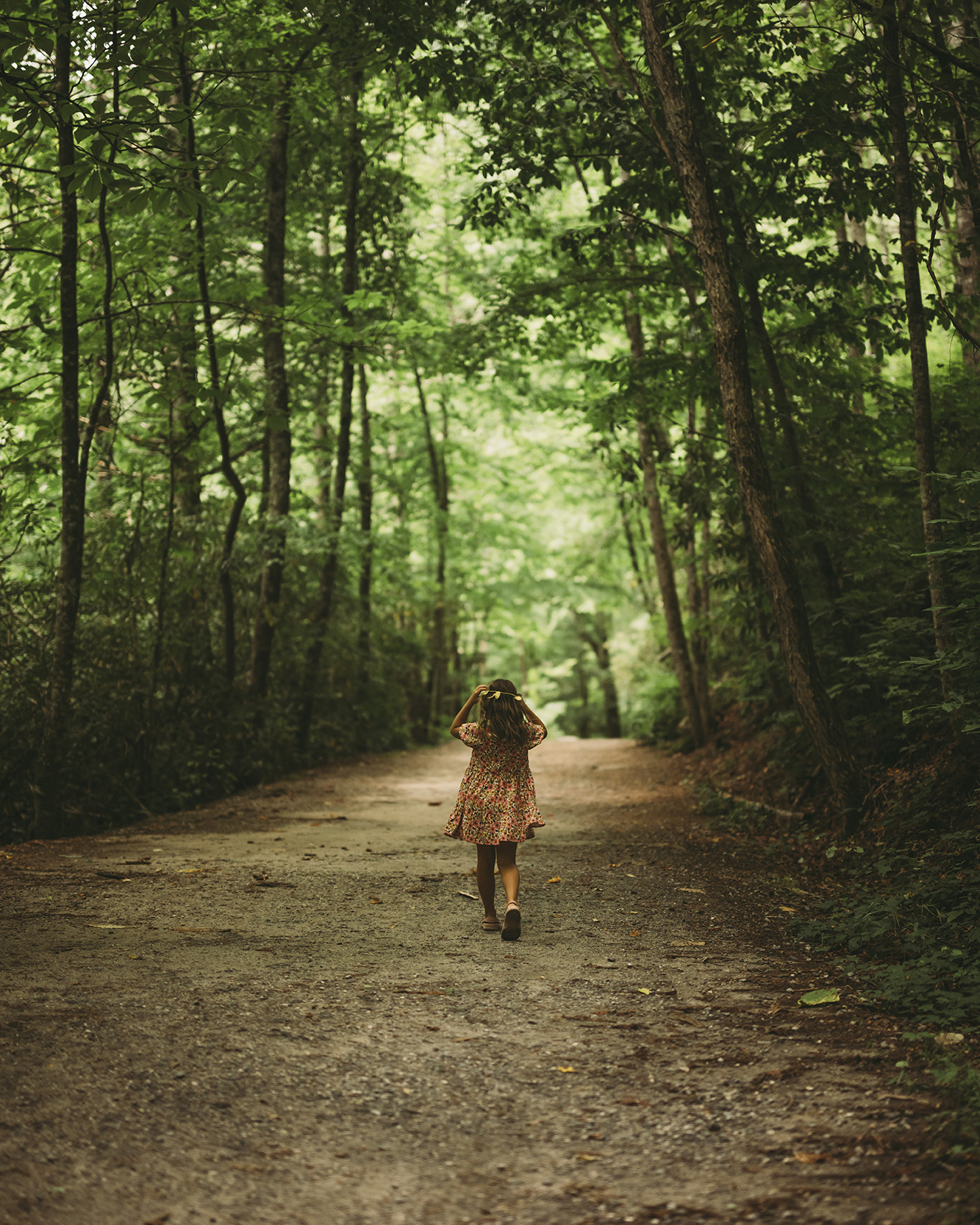 Girl in a floral dress walks away along a dirt forest path framed by tall trees.