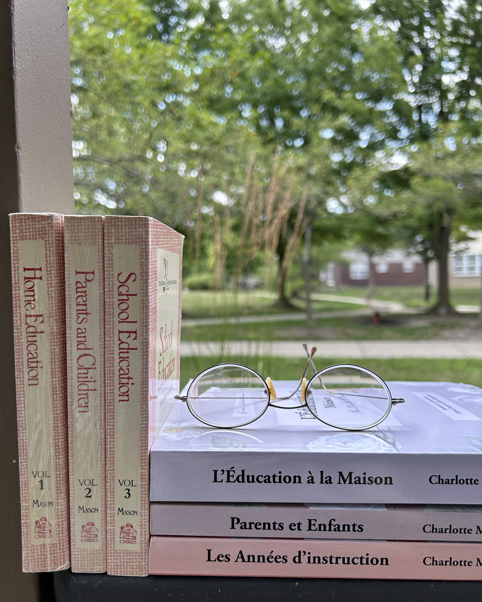 Stack of pink home-education books on a table with a pair of round glasses, outdoor greenery blurred in the background.
