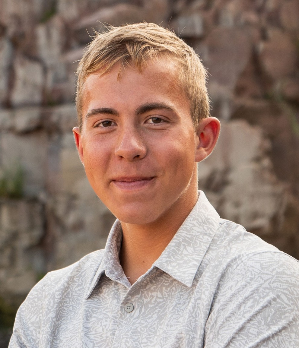 Young person with short blonde hair, wearing a light patterned button-down shirt, smiling softly outdoors with a rocky background