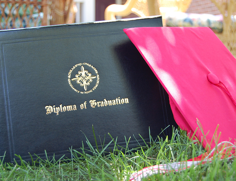 Diploma cover with a gold crest and the words 'Diploma of Graduation' lying on grass next to a pink graduation cap outdoors.