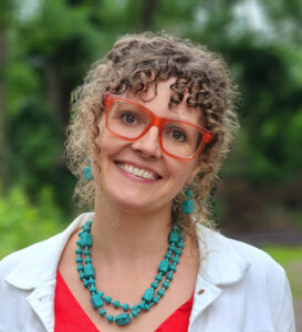 Smiling woman with curly hair wearing red glasses and turquoise beaded necklace outdoors.