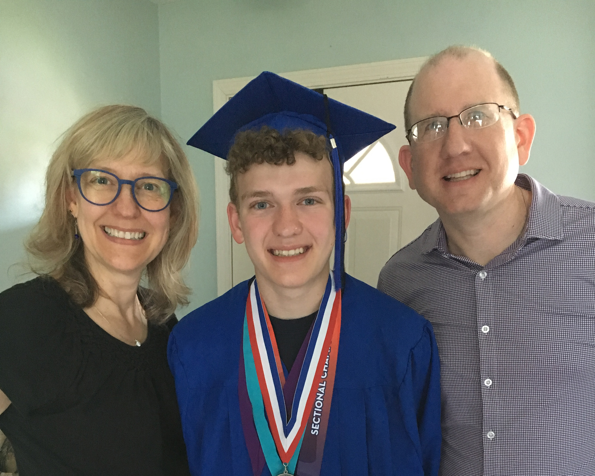 Graduate in a blue cap and gown smiling, flanked by two family members in a doorway. The graduate wears red-white-blue medals around the neck.